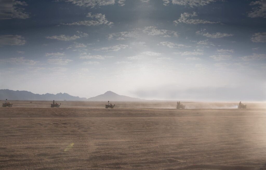 Quad bike convoy racing across the endless golden sands of Sharm El Sheikh's Sinai desert, with hazy mountains on the horizon under a dramatic cloudy sky