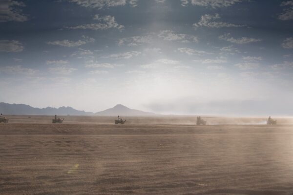 Quad bike convoy racing across the endless golden sands of Sharm El Sheikh's Sinai desert, with hazy mountains on the horizon under a dramatic cloudy sky