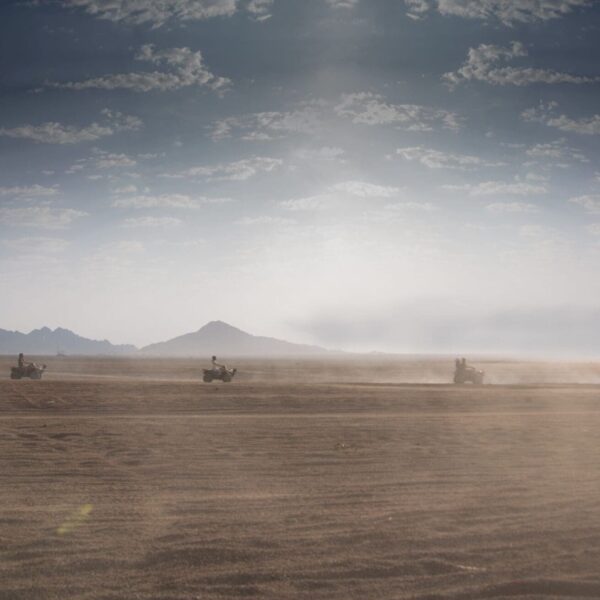 Quad bike convoy racing across the endless golden sands of Sharm El Sheikh's Sinai desert, with hazy mountains on the horizon under a dramatic cloudy sky