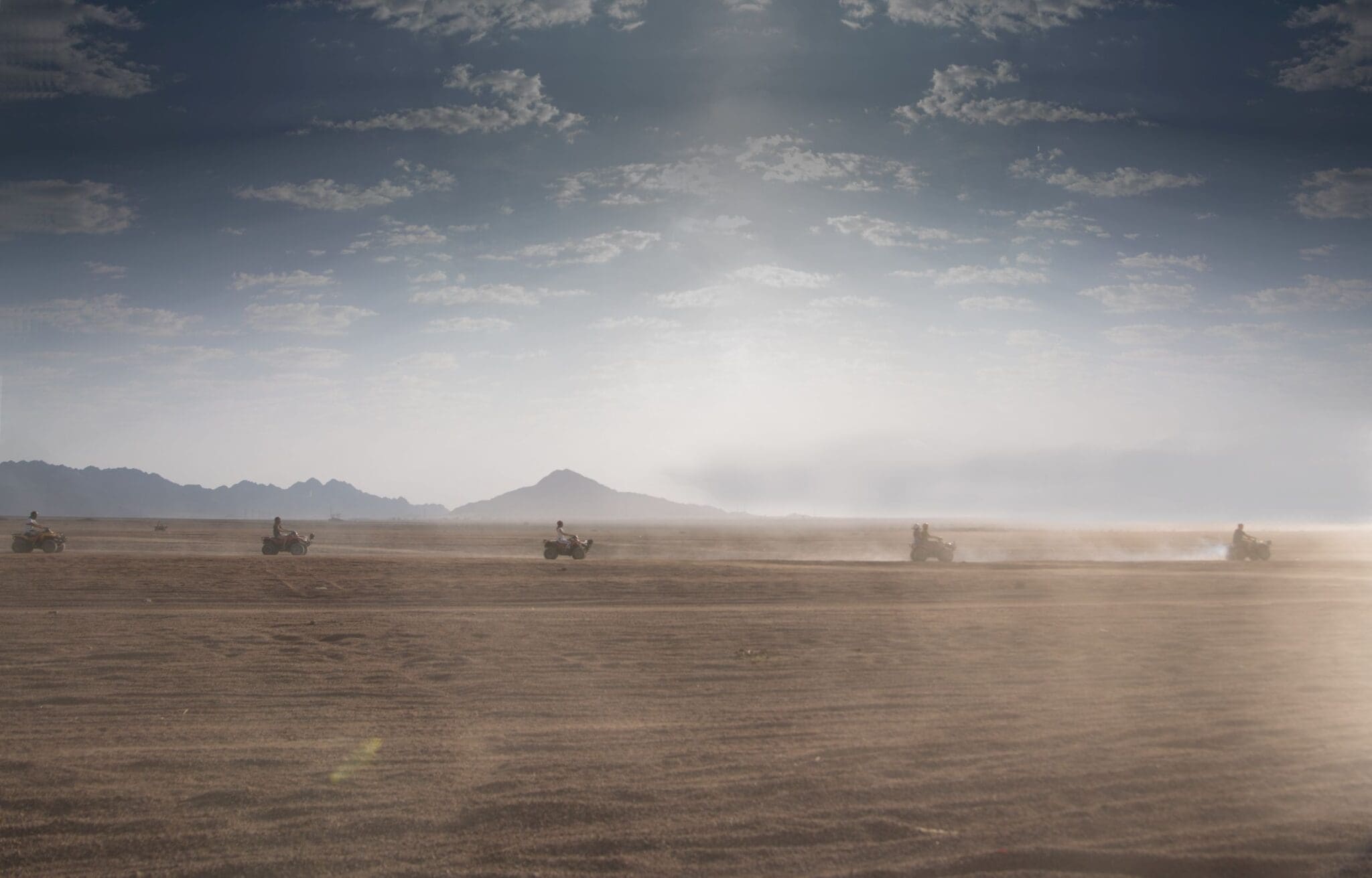 Quad bike convoy racing across the endless golden sands of Sharm El Sheikh's Sinai desert, with hazy mountains on the horizon under a dramatic cloudy sky